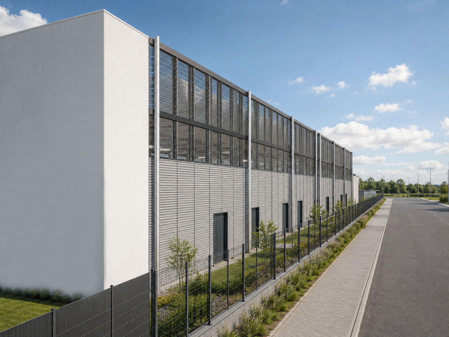 Seitenansicht eines Rechenzentrums mit grauer Lamellenfassade, weißen Wänden, Sicherheitszaun und Grünstreifen an der Straße.